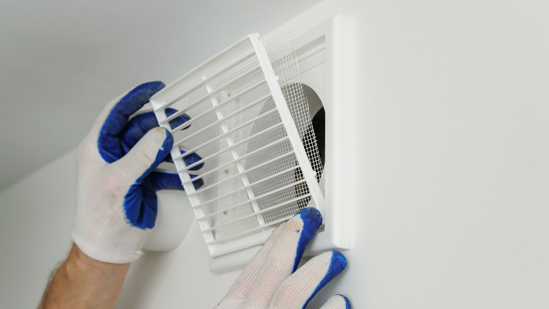 Gloved hands installing a white positive input ventilation (PIV) unit on a white ceiling to improve indoor air circulation.