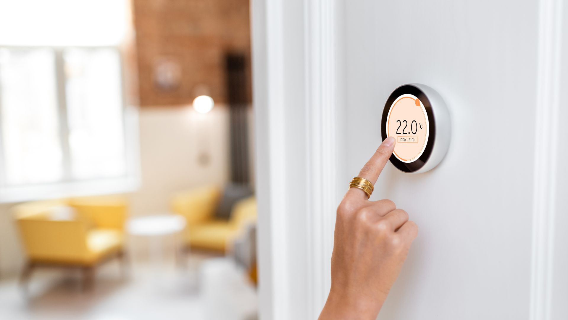 Hand adjusting a digital thermostat on a white wall, set to 22°C, with a modern living room in the background.