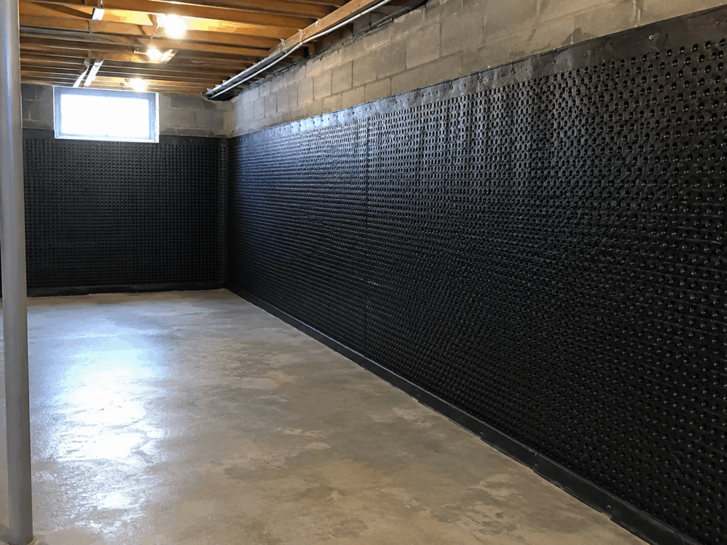 Residential basement waterproofing system installed on interior walls, showing black cavity drain membrane fixed to concrete block walls, exposed timber ceiling joists, a small basement window, and a clean concrete floor ready for renovation.